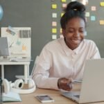 Emprego Remoto com R$ 1.200 de VA/VR Woman smiling while working on a laptop at a desk.