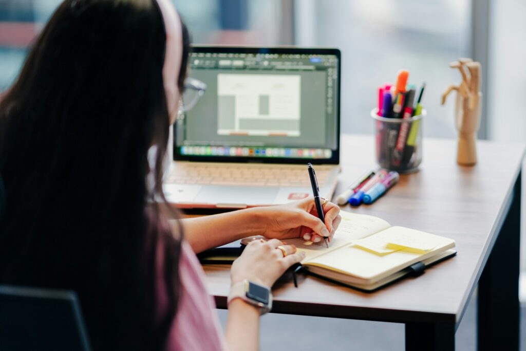 Vagas Remotas na Enjoei, com VR de R$ 900 e mais benefícios! Woman working at a desk with laptop and notebook.