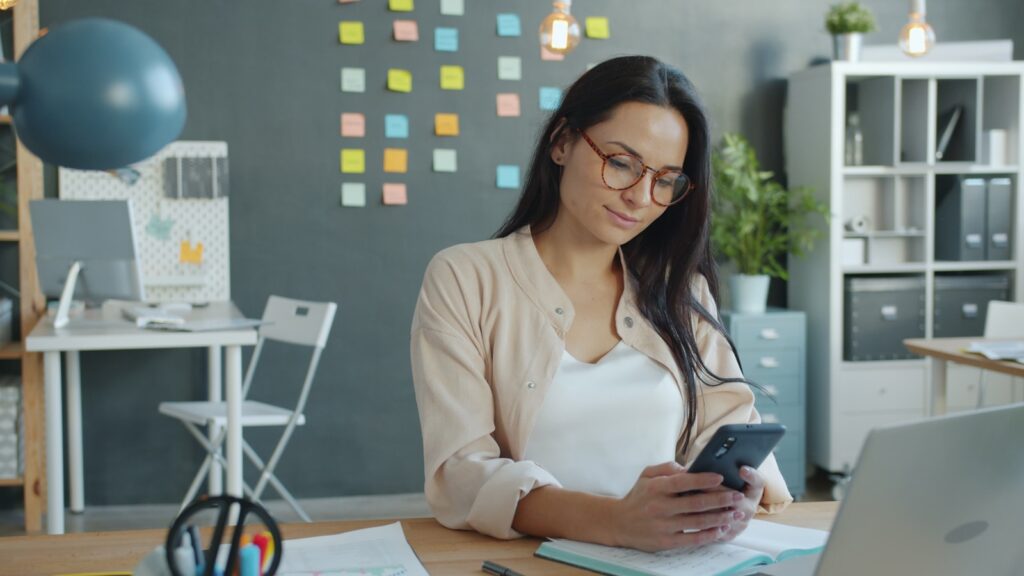 Vaga Remota com Salário de R$ 3.600,00 Woman looking at her phone in an office.