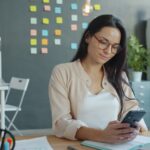 Vaga Remota com Salário de R$ 3.600,00 Woman looking at her phone in an office.