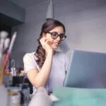 Sucesso do Cliente JR e + remotas com VA de R$ 718,00 + comissões! A cheerful woman in glasses works remotely on her laptop in a cozy home office setting.