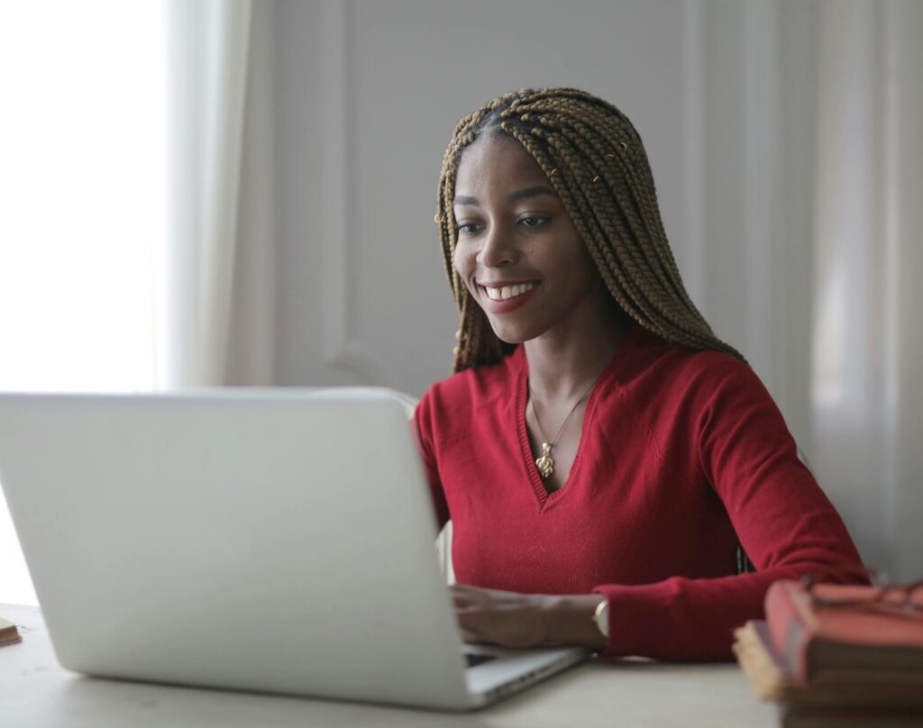 Vagas Remotas na VExpenses, VA/VR de R$ 1.200 Young black woman happily working on a laptop indoors, showcasing a modern remote work setup.