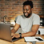 Remotas com salário acima de R$ 7.500 (CLT) African American man smiling while working remotely on laptop from home office