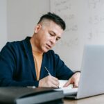 Auxiliar Administrativo (Remoto) e + com VA/VR até R$ 1.500! Focused professional writing notes at home office desk with laptop.