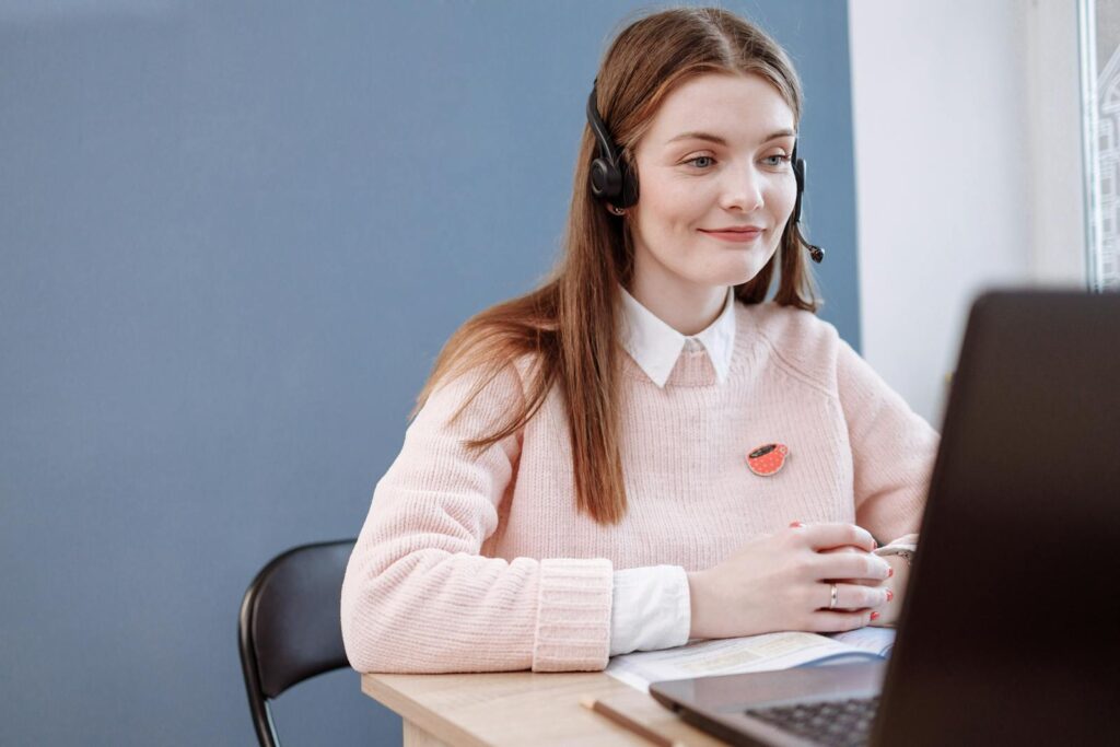 Sucesso do Cliente JR e + remotas com VA de R$ 718,00 + comissões! Smiling woman in pink sweater working remotely as a customer service agent.