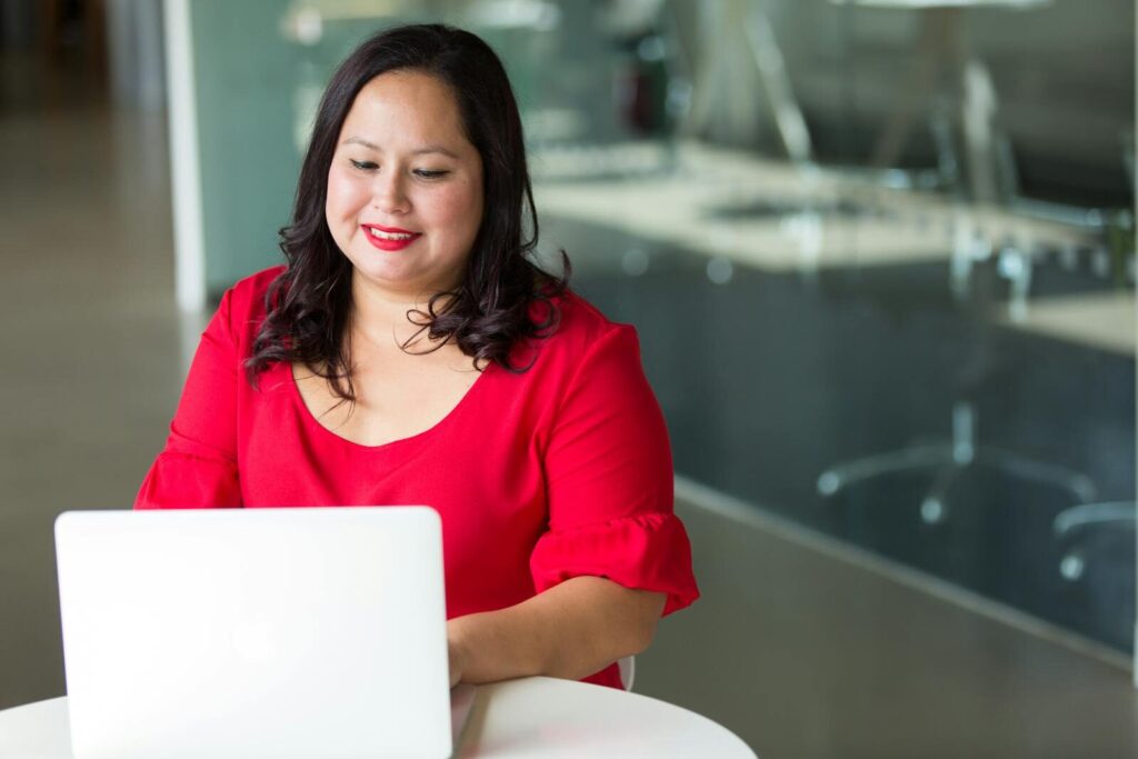 Vagas Remotas na Vox, com VA/VR de R$ 750,00 Asian woman smiling while working on a laptop in a modern office environment.