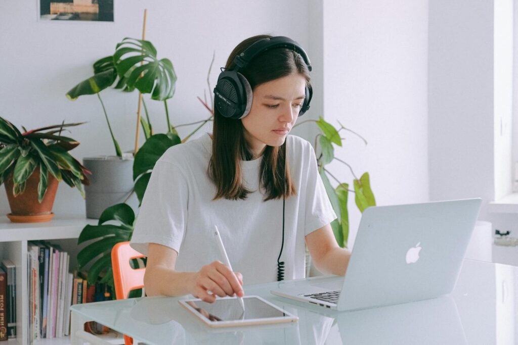 Analista Administrativo (Remoto) e + com VR/VA até R$ 1.500! Young woman in home office using a laptop and drawing tablet, wearing headphones, illustrating remote work.