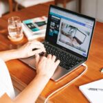 Home Office na Pulsus, com VA/VR de R$ 900,00 A person working remotely on a laptop at a wooden desk with stationery and a notepad.