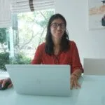 Vagas Remotas na Minsait Woman works on laptop at a bright, modern table.