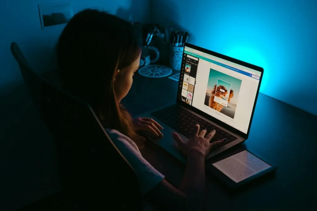 Teletrabalho com VA/VR de R$ 1.000,00 A girl uses a laptop at a desk in a dimly lit room, with a blue light creating a calm ambiance.