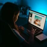 Teletrabalho com VA/VR de R$ 1.000,00 A girl uses a laptop at a desk in a dimly lit room, with a blue light creating a calm ambiance.