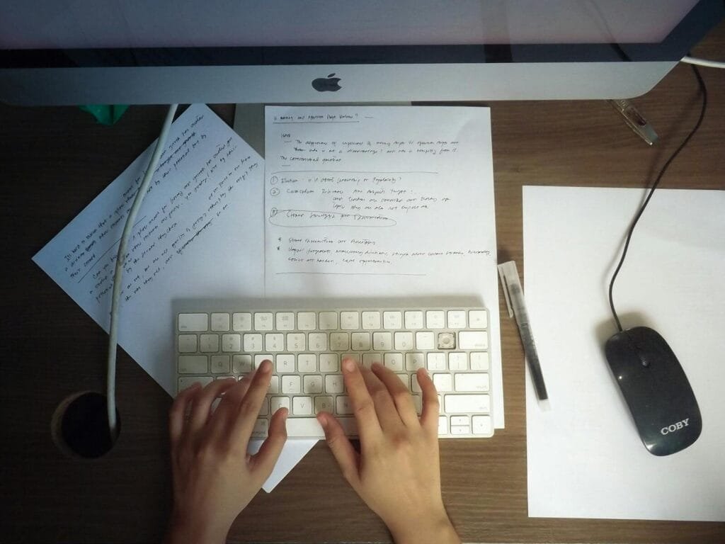 Assistente de Atendimento Home Office e +vagas Hands typing on a keyboard at a wooden desk with papers and computer mouse visible.