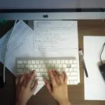 Home Office com salário até R$ 6.000 (CLT) Hands typing on a keyboard at a wooden desk with papers and computer mouse visible.