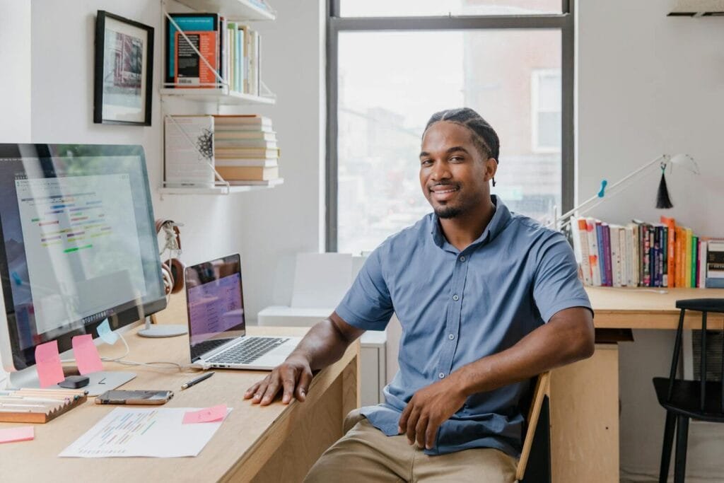 Unicred contratando para Home Office com R$ 1.000,00 de VA/VR Confident professional man with a welcoming smile sitting at a desk in a contemporary office setting.
