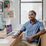 Unicred contratando para Home Office com R$ 1.000,00 de VA/VR Confident professional man with a welcoming smile sitting at a desk in a contemporary office setting.