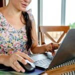 Neogrid contratando para Home Office A woman using a laptop for remote work indoors, featuring a floral dress and henna design.