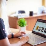 BRQ contrata para Home Office, com VA/VR de R$ 1.950,00 A woman browsing an online store on a laptop in a modern home office setting.