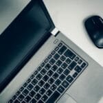 Assistente Administrativo e mais vagas remotas A close-up view of a silver laptop and wireless mouse on a smooth table, ideal for home office setups.
