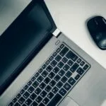 Assistente Administrativo e mais vagas remotas A close-up view of a silver laptop and wireless mouse on a smooth table, ideal for home office setups.