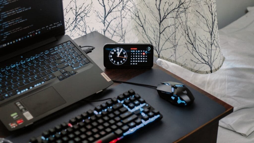 Assistente de Atendimento e +Remotas com VA/VR de R$ 950,00 A modern workspace featuring a laptop, digital clock, gaming mouse, and keyboard, ideal for work and tech enthusiasts.