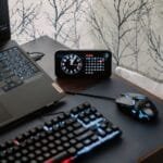 Assistente de Atendimento e +Remotas com VA/VR de R$ 950,00 A modern workspace featuring a laptop, digital clock, gaming mouse, and keyboard, ideal for work and tech enthusiasts.