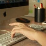 Home Office na Linx, com VA/VR de R$ 800,00 A close-up of a woman's hand typing on an Apple keyboard in an office setting.