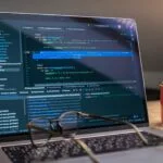 Vagas Remotas na Nexdom, com VA/VR de R$ 950,00 A laptop displaying code editor with a motivational mug that reads 'Make It Happen' on a workspace.