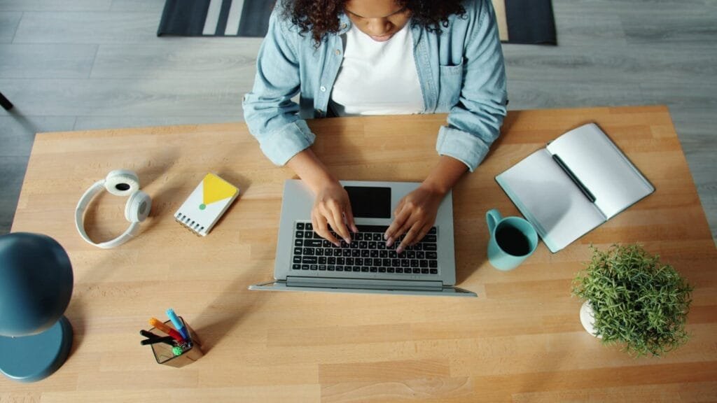 Analista Administrativo e + Remotas com VR/VA até R$ 1.500! A woman using a laptop at a wooden desk with headphones, a notebook, and a plant.