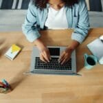 Analista Administrativo e + Remotas com VR/VA até R$ 1.500! A woman using a laptop at a wooden desk with headphones, a notebook, and a plant.