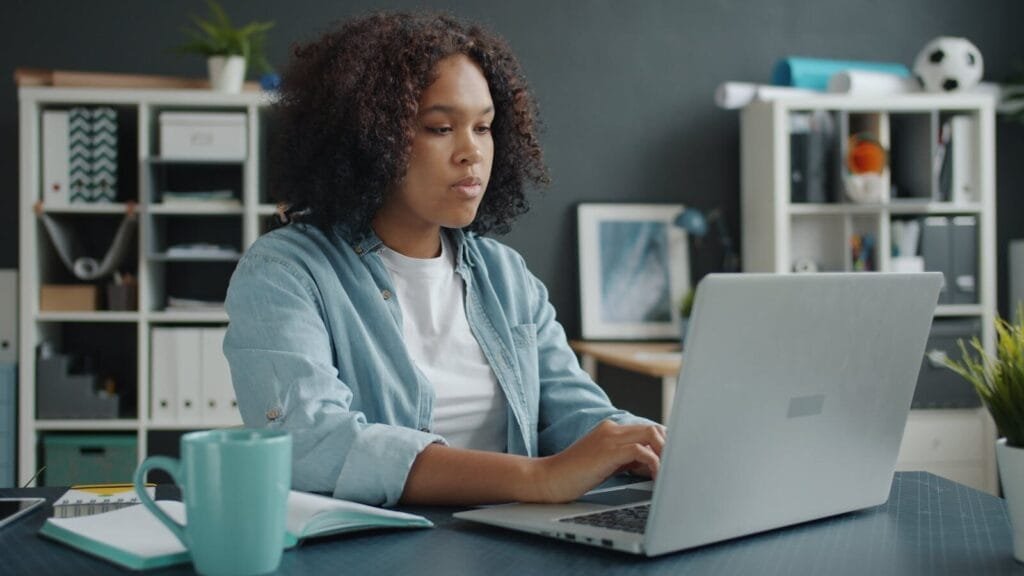 Web Estratégica contratando para vagas 100% remotas! Young woman focused on remote work at home office desk with laptop.