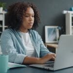 Web Estratégica contratando para vagas 100% remotas! Young woman focused on remote work at home office desk with laptop.