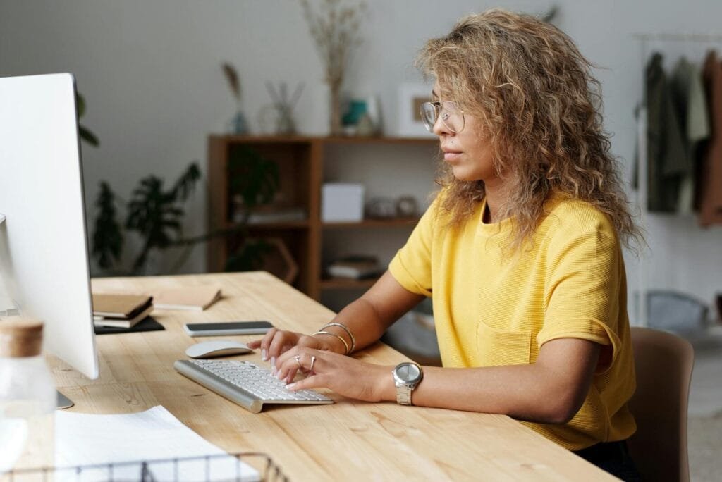 Assistente de Prevenção à Fraude e +vagas home office A woman working remotely from home, typing on a keyboard at her work desk.
