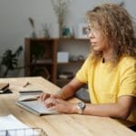 Assistente de Prevenção à Fraude e +vagas home office A woman working remotely from home, typing on a keyboard at her work desk.