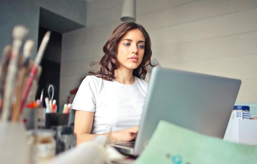 Confitec contratando para Vagas Remotas, R$ 800 de VA/VR A woman concentrating on her laptop in a creative indoor setting, surrounded by art supplies.