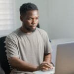 Vagas remotas para Assistente Administrativo e + African American man working remotely on a laptop in a well-lit indoor setting.