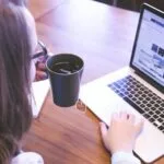 Vinta contrata Home Office com R$ 1.600 de VA/VR e salário até 18.300! A woman enjoys coffee while browsing the web on a laptop at a home office desk.