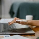 Analista de Gestão e mais vagas remotas! A woman works remotely on her laptop while holding a pen, focused on her task in a casual home setting.