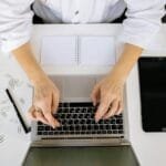 Join Tecnologia contratando para 100% Home Office A workspace with a laptop, digital tablet, and hands typing, viewed from above.