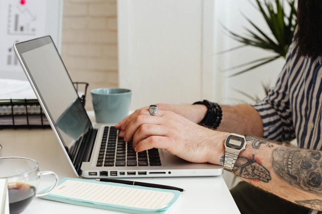 Emprego remoto: vagas para Assistente de Suporte e mais áreas A person with tattoos types on a laptop at a desk, sipping coffee and taking notes.