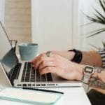 Emprego remoto: vagas para Assistente de Suporte e mais áreas A person with tattoos types on a laptop at a desk, sipping coffee and taking notes.