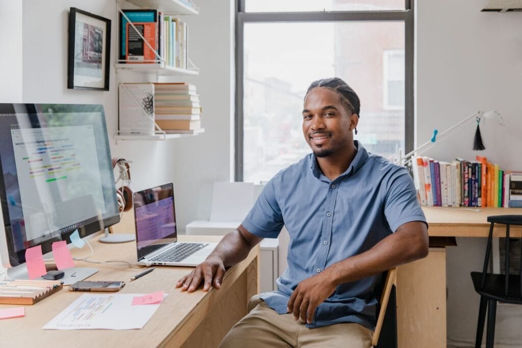 Sucesso do Cliente e +vagas com R$ 1.012,50 de VA/VR Confident professional man with a welcoming smile sitting at a desk in a contemporary office setting.