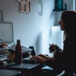 Sucesso do Cliente e +vagas com R$ 1.012,50 de VA/VR Silhouette of a woman working at her desk with a cat nearby in a cozy home office setup.