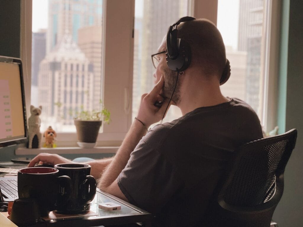 Hesselbach contratando para Home Office A man wearing headphones works at his desk overlooking a city skyline from a home office.