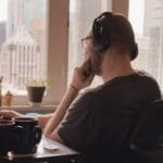 Hesselbach contratando para Home Office A man wearing headphones works at his desk overlooking a city skyline from a home office.