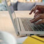 Desenvolvedor Cobol e mais Vagas 100% Remotas! Hands typing on a laptop keyboard indoors with blurred background and natural lighting.
