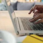 Desenvolvedor Cobol e mais Vagas 100% Remotas! Hands typing on a laptop keyboard indoors with blurred background and natural lighting.