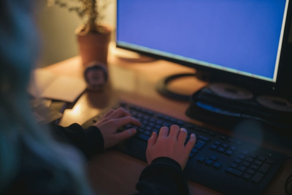 Analista de BI Júnior e mais Home Office A woman typing on a keyboard in a dimly lit, cozy workspace, featuring a computer screen.