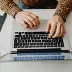 Vagas Remotas com R$ 932 de VR/VA Hands typing on a laptop keyboard in a modern home office environment.