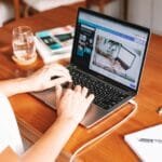 Home Office para Assistente Comercial e mais! A person working remotely on a laptop at a wooden desk with stationery and a notepad.