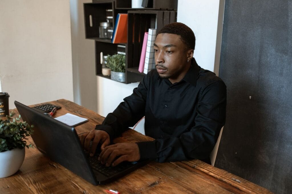 Emprego Remoto com salário a partir de R$ 16.000 (CLT) African American man in black shirt typing on laptop at office desk